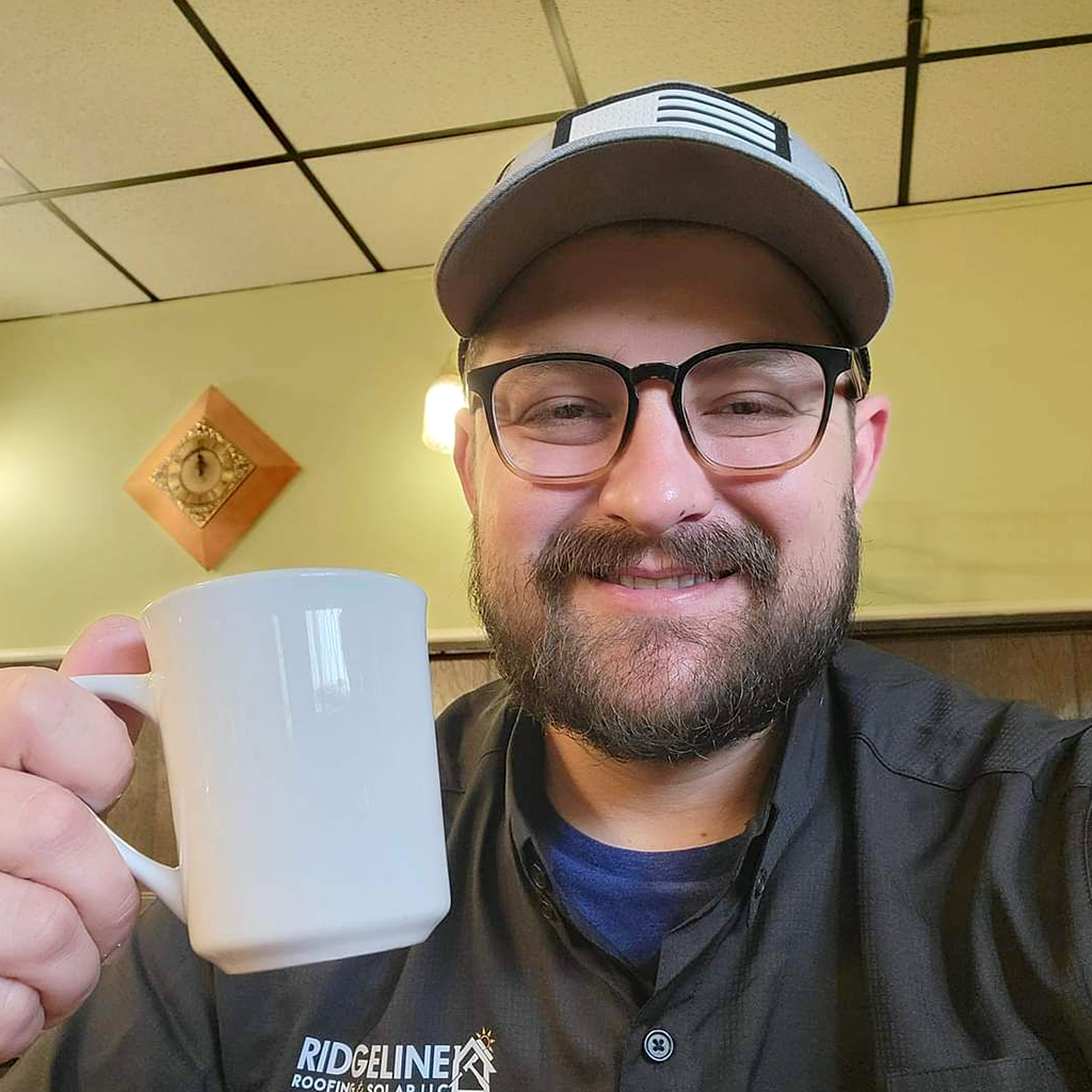 Stephen Walker, Project Manager with Ridgeline Roofing and Solar of Joplin, MO, is holding a white coffee cup and smiling while sitting in a diner planning a new roof installation.